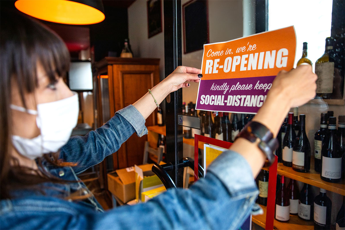Woman wearing a mask and hanging an orange and purple sign that says "Come in, we're reopening. Kindly please keep social-distancing"