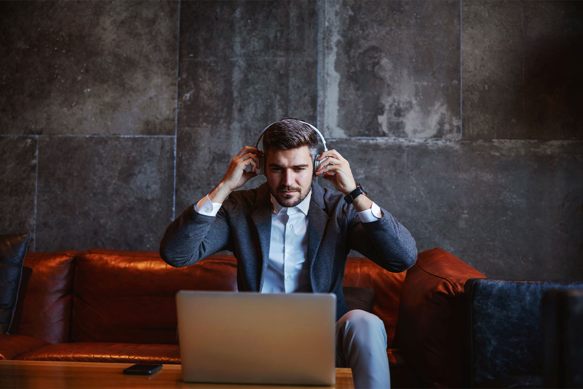 A man sitting on a couch looking at his laptop on a coffee table in front of him while putting headphones over his ears.